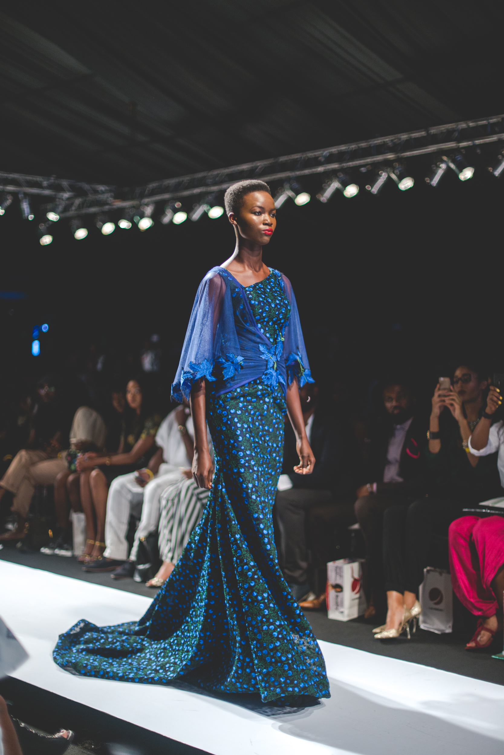 A model walking the runway in a blue patterned gown at Lagos Fashion and Design Week, with photographers and guests seated alongside